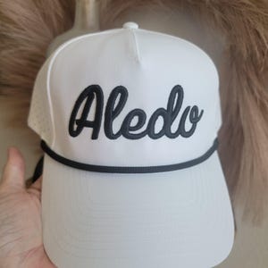 May include: White baseball cap with the word "Aledo" embroidered in black script. The cap features a black rope detail and a perforated side panel for ventilation. A classic and stylish accessory.