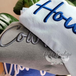 May include: Three t-shirts stacked on top of each other. The top shirt is light blue with the word "Howe" embroidered in blue. The middle shirt is gray with the word "Cole" embroidered in gray. The bottom shirt is blue with the word "You" embroidered in white. The shirts are all folded and stacked on top of each other.