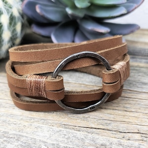 May include: A brown leather wrap bracelet with a silver-colored metal ring clasp. The bracelet has multiple leather straps and is stitched with tan thread. The bracelet is shown on a wooden surface.