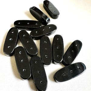 May include: A collection of glossy black oval-shaped beads, each with two small holes. The beads are scattered across a white surface, with varying angles and orientations, creating a visually appealing arrangement.