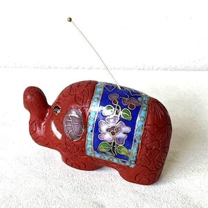 May include: A red cloisonné elephant figurine with blue, white, and green floral designs. The elephant has a gold antenna on its head.