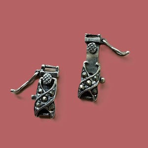 May include: Two silver-toned jewelry clasps with intricate detailing. Each clasp features a lever and is adorned with small, dark gemstones and decorative patterns. The clasps are set against a muted red-brown background.