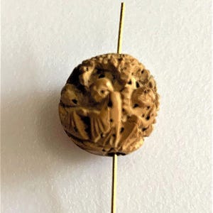 May include: A small, intricately carved wooden bead with a detailed relief depicting figures. The bead is a light brown color and is pierced by a thin, gold-colored metal rod. The carving shows multiple figures.