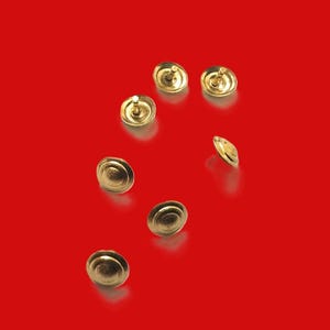 May include: Seven gold-colored upholstery tacks are scattered on a red background. Each tack has a rounded head and a central pin. The tacks are evenly spaced, with some oriented at different angles, creating a sense of depth.