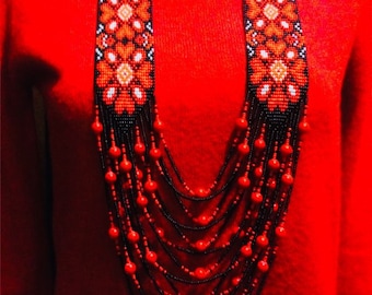 May include: A multi-strand beaded necklace with red and black beads. The necklace features a beaded centerpiece with a floral pattern in red, white, and black.