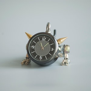 May include: A black clock with gold accents and a black face with white numbers. The clock is attached to a metal base with two gold spikes.