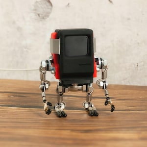 May include: A small robot sculpture with a black rectangular head and a red accent. The robot's body is constructed from metal hardware, including bolts, nuts, and hinges. The robot has articulated arms and legs, and stands on a wooden surface.