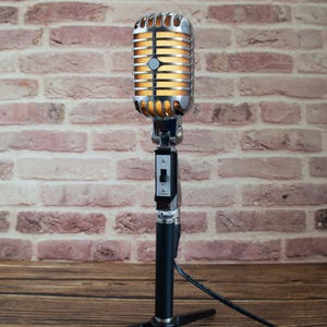 May include: A vintage-style microphone with a silver finish and a black stand. The microphone has a large, round head with a grill and a yellow-orange light inside.