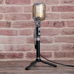 May include: A vintage-style silver microphone with a ribbed metal head and a black cylindrical body. The microphone features a black toggle switch on the front and is mounted on a black three-legged tripod stand with a black cable extending to the right.