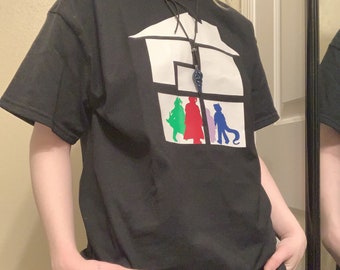 May include: Black t-shirt with a white graphic of a house with four figures inside. The figures are wearing different colored clothing and are standing in a row. The house graphic has a black cord with a pendant hanging from it.