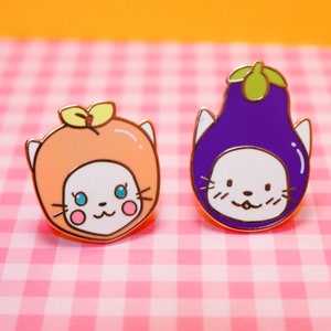 May include: Two enamel pins featuring cartoon cats wearing fruit and vegetable costumes. The first pin is a white cat wearing an orange peach costume. The second pin is a white cat wearing a purple aubergine costume.