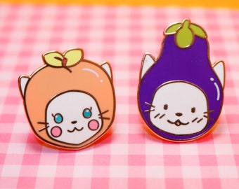 May include: Two enamel pins featuring cartoon cats wearing fruit and vegetable costumes. The first pin is a white cat wearing an orange peach costume. The second pin is a white cat wearing a purple eggplant costume.