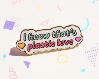 May include: A white enamel pin with a gold border and the text "I know that's plastic love" in blue and pink lettering. There are two small pink hearts on either side of the text.