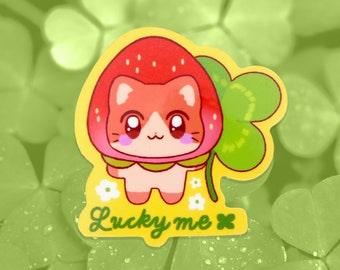 May include: A cute cartoon cat wearing a red strawberry hat and holding a green four-leaf clover. The cat is standing on a yellow background with white flowers and the text "Lucky me" written below.
