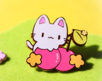 May include: White cat with pink cheeks inside a pink cherry with a green leaf and a yellow star. The cat is wearing a pink cherry costume.