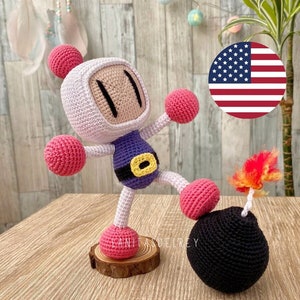 Puede incluir: Un juguete de peluche tejido a crochet de un personaje de videojuego, Bomberman, de pie sobre una base de madera. Lleva un atuendo morado y amarillo y sostiene una bomba negra con una mecha roja y naranja. El juguete está frente a un fondo de madera con una bandera estadounidense en el fondo.