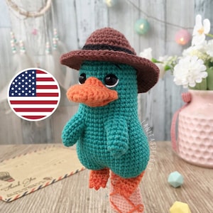 Puede incluir: Un juguete de peluche de ganchillo de un ornitorrinco con un sombrero marrón. El ornitorrinco es verde azulado con patas naranjas y una cola rosa. El juguete está de pie sobre una rodaja de madera.