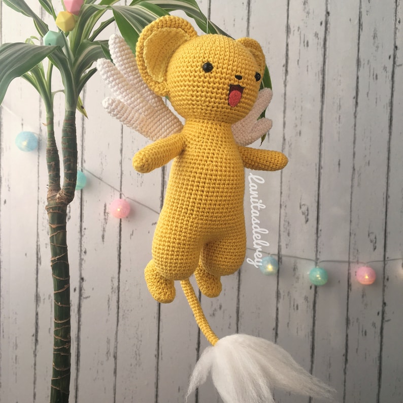 Puede incluir: Un juguete de peluche amarillo tejido a crochet con alas blancas y una cola blanca esponjosa. El juguete tiene una cara de dibujos animados con ojos negros y una lengua rosa que sobresale.