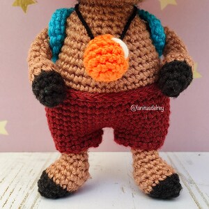 Puede incluir: Juguete amigurumi de crochet con pelaje marr&oacute;n, vistiendo una camisa a rayas rojas y blancas, una mochila azul y un collar naranja y blanco. El juguete tiene pies y manos negros.