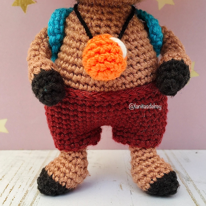 Puede incluir: Juguete amigurumi de crochet con pelaje marr&oacute;n, vistiendo una camisa a rayas rojas y blancas, una mochila azul y un collar naranja y blanco. El juguete tiene pies y manos negros.