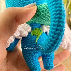 May include: A handmade blue crochet character with white wings and a green leaf emblem. The character has a green patch on its back and a green and white patch on its head. The text "Lanitas del Rey" is visible on the bottom of the character.
