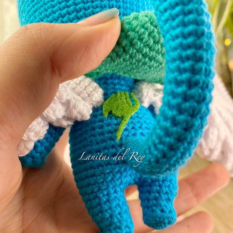 May include: A handmade blue crochet character with white wings and a green leaf emblem. The character has a green patch on its back and a green and white patch on its head. The text "Lanitas del Rey" is visible on the bottom of the character.