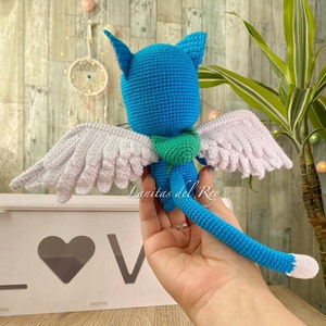 May include: A handmade blue crochet cat with white wings and a green scarf. The cat has a long tail and pointed ears. The wings are detailed with individual feathers. The text "Lanitas del Rey" is visible.