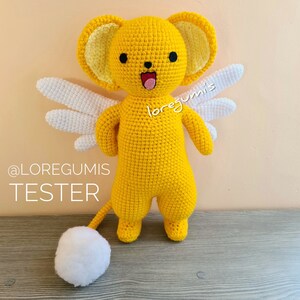 Puede incluir: Un animal de peluche amarillo tejido a crochet con alas blancas y una cola blanca. El animal tiene una cara sonriente con ojos negros y una lengua rosa. El animal est&aacute; de pie sobre una superficie de madera. El texto "@LORE GUMIS TESTER" es visible en la imagen.