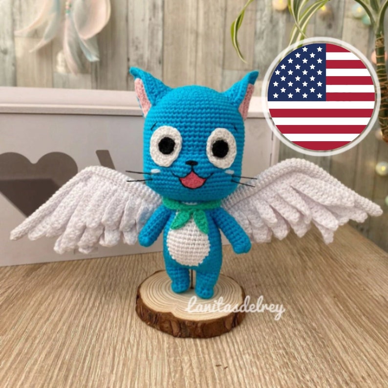 May include: A handmade crochet cat figurine with blue body, white wings, and a green scarf. The cat has large white eyes, pink inner ears, and a smiling pink mouth. It stands on a small wooden base. The figurine is a whimsical, handcrafted item.