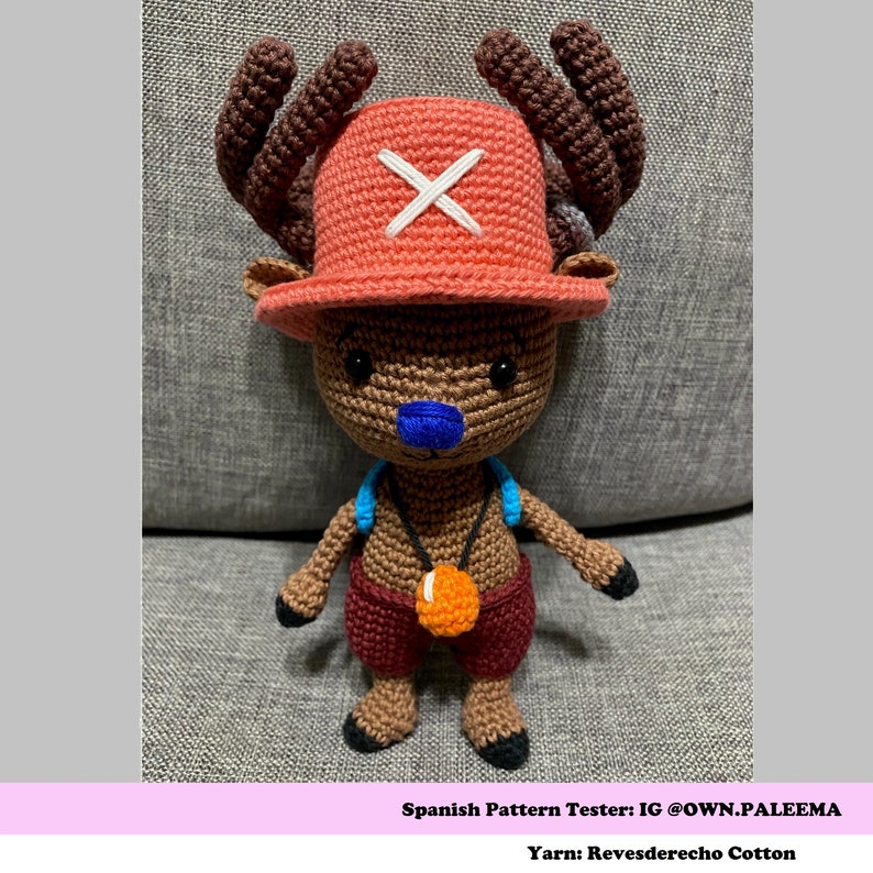 Puede incluir: Juguete de reno de crochet con pelaje marr&oacute;n, pantalones cortos rojos, un chaleco azul y un sombrero rojo con una X blanca. El reno sostiene una bola naranja en una cuerda.