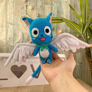 May include: A handmade blue crochet cat with white wings, a green scarf, and a pink tongue. The cat has large black and white eyes and black whiskers. The wings are white and detailed. The text "Lanitas del Rey" is visible.