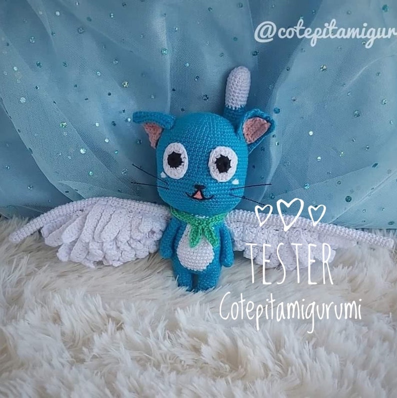 May include: A blue crochet cat with white wings, a green scarf, and large black eyes. The cat has a white tail and pink inner ears. The text "TESTER" and "Cotepitamiurumi" are visible. The background is a fluffy white surface and blue fabric.
