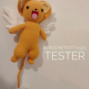 Puede incluir: Un juguete de peluche amarillo y blanco tejido a crochet con una cola larga, alas y una cara sonriente. El juguete est&aacute; colgado del techo y el texto "@CROCHETKITTY1323 TESTER" es visible debajo del juguete.