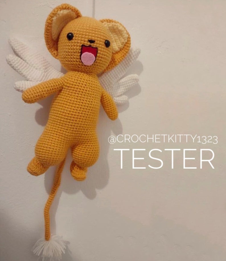 Puede incluir: Un juguete de peluche amarillo y blanco tejido a crochet con una cola larga, alas y una cara sonriente. El juguete est&aacute; colgado del techo y el texto "@CROCHETKITTY1323 TESTER" es visible debajo del juguete.