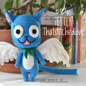May include: A handmade blue crochet cat with white wings, a green scarf, and a white belly. The cat has large white eyes with black pupils and a pink tongue. The text "TESTER Thatstitchedlove" is written on the image.