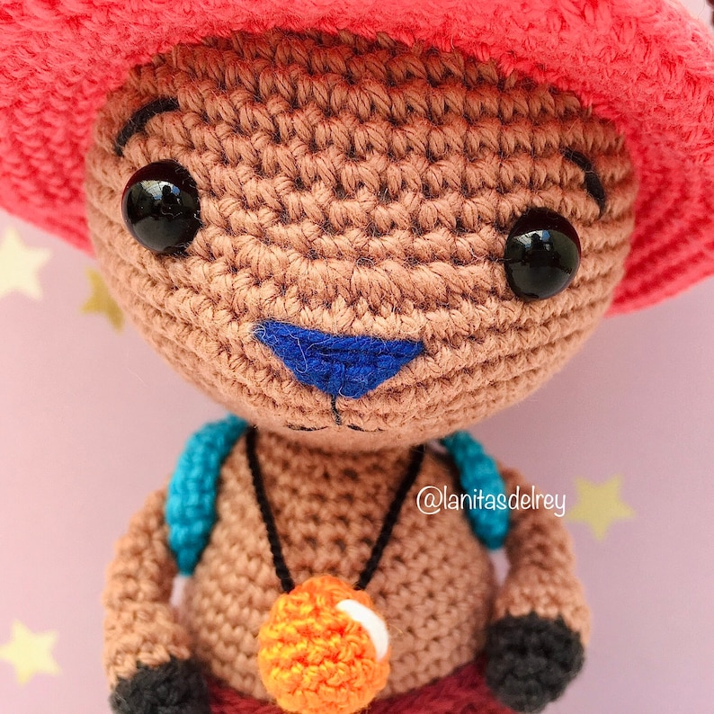 Puede incluir: Juguete de amigurumi tejido a crochet de un personaje que lleva un sombrero rojo, una camisa azul y un collar con un colgante naranja. El juguete tiene hilo marr&oacute;n para el cuerpo e hilo negro para los ojos.