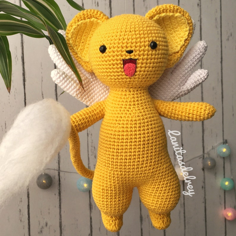 Puede incluir: Un juguete de peluche amarillo tejido a crochet con alas blancas y cola. El juguete tiene una cara de dibujos animados con ojos negros, lengua rosa y nariz negra. El juguete est&aacute; colgado de una cuerda.