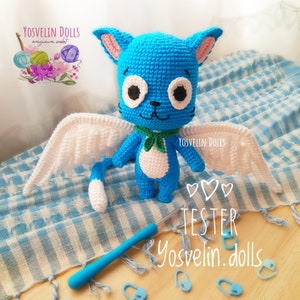 May include: A handmade blue crochet cat doll with white wings, a green scarf, and a white belly. The doll has large white eyes with black pupils and pink-lined ears. The image includes a crochet hook, stitch markers, and the text "Yosvelin Dolls".