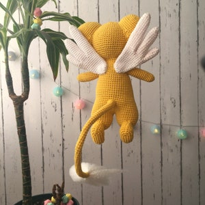 Puede incluir: Un juguete de peluche amarillo tejido a crochet con alas blancas y una cola larga. El juguete est&aacute; colgado del techo.
