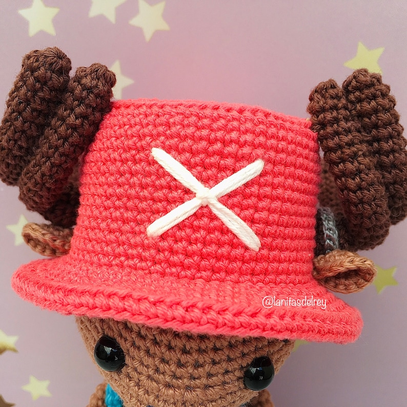 Puede incluir: Juguete de peluche tejido a crochet con cuernos marrones y un sombrero rojo con una X blanca en la parte delantera. El sombrero tiene un borde y el juguete tiene ojos negros.