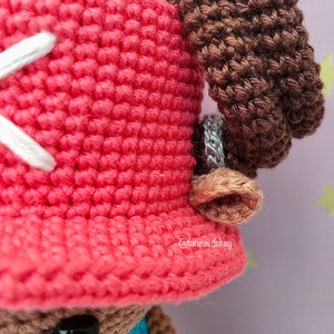 Puede incluir: Primer plano de la cabeza de una mu&ntilde;eca de crochet con un sombrero rojo y cabello casta&ntilde;o. El sombrero tiene una X blanca cosida en &eacute;l. La oreja de la mu&ntilde;eca es visible en el lado derecho de la imagen. El sombrero de la mu&ntilde;eca tiene el texto "@lanitasdelrey" en el borde.