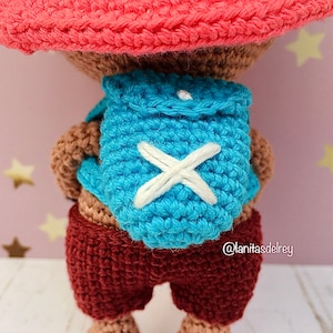 Puede incluir: Un juguete de crochet que lleva un sombrero rojo y una mochila azul con una X blanca en la parte delantera. El juguete lleva pantalones marrones.
