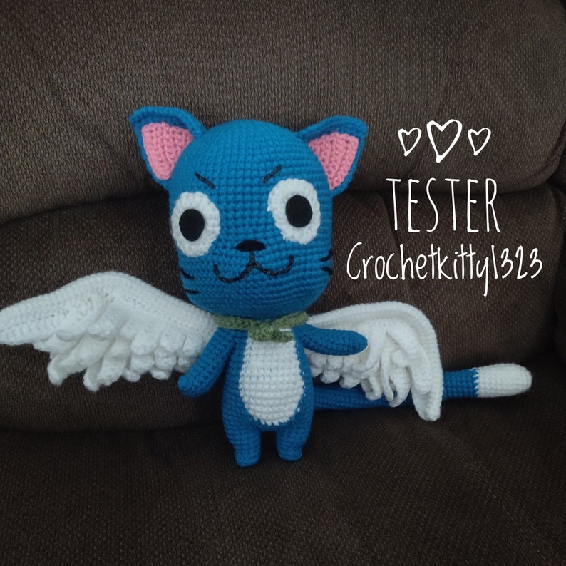 May include: A handmade blue crochet cat with white wings, pink ears, and a green collar. The cat has large white eyes with black pupils and a smiling face. The text "TESTER Crochetkitty1323" is written on the image.