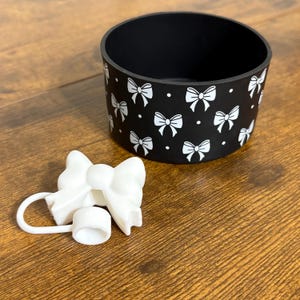 May include: A black cup holder with a white bow pattern and a white bow-shaped lid. The cup holder is cylindrical and sits on a wooden surface. The lid is attached to a small white cap.