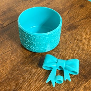 May include: A turquoise container with a bow pattern and a matching bow and ring. The container is cylindrical with a repeating bow design around the sides. The bow and ring are separate pieces.