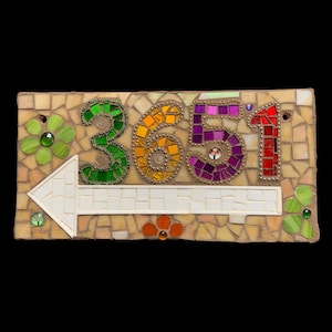 Puede incluir: Un letrero de dirección de mosaico con el número 3651 y una flecha. Los números son verdes, amarillos, morados y rojos. El letrero tiene un fondo beige con mosaicos y detalles de vidrio decorativos.