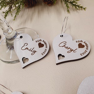 May include: Two white wooden heart-shaped wine glass charms with the names "Emily" and "Cheryl" engraved on them. The charms also include the date "25th August 2023" and a small heart.