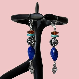 May include: A pair of dangle earrings with a combination of blue, turquoise, and amber-colored beads. The earrings feature a silver filigree bead and a diamond-shaped charm. The earrings are displayed on a black stand against a pink background.