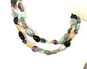 May include: A two-strand necklace featuring a variety of colorful, polished gemstones. The stones include shades of purple, green, blue, black, and yellow. The necklace is displayed on a white background.