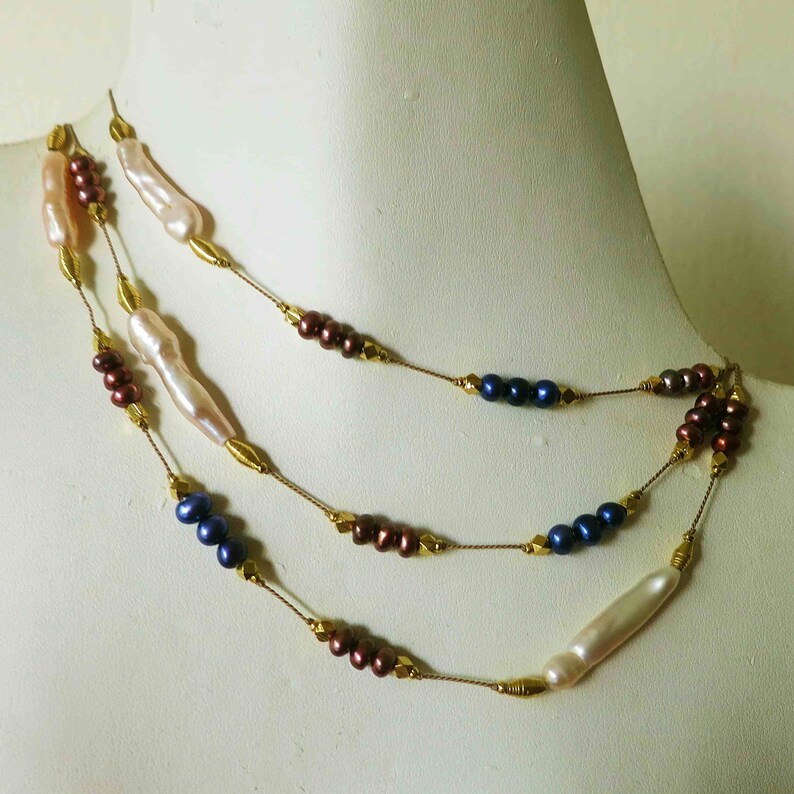 May include: A multi-strand necklace with gold chain and pearls. The necklace features three strands of pearls in different colors: white, blue, and brown.